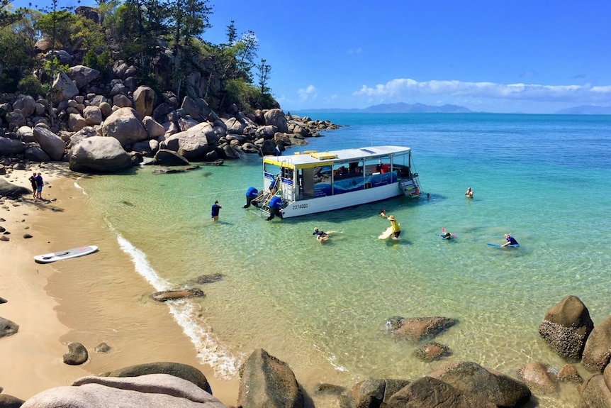 Vista de pájaro de un pequeño barco en una playa desierta en la Isla Magnética, nadadores nadando alrededor del barco en aguas claras.