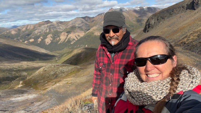 Un hombre con una camisa a cuadros roja y una gorra de béisbol y una mujer con una gran bufanda gris están uno al lado del otro contra un fondo de montañas.