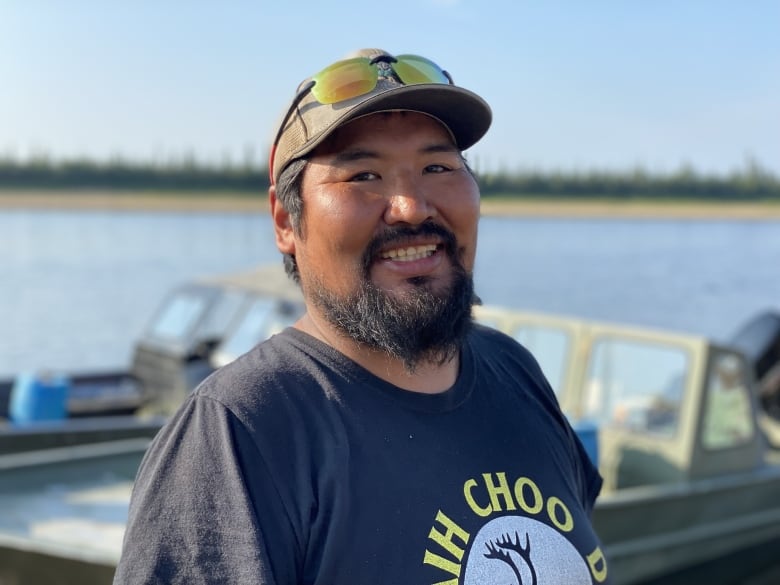 Un hombre con gorra de béisbol y gafas de sol mira a la cámara y sonríe, con un río azul y un barco de pesca detrás de él.