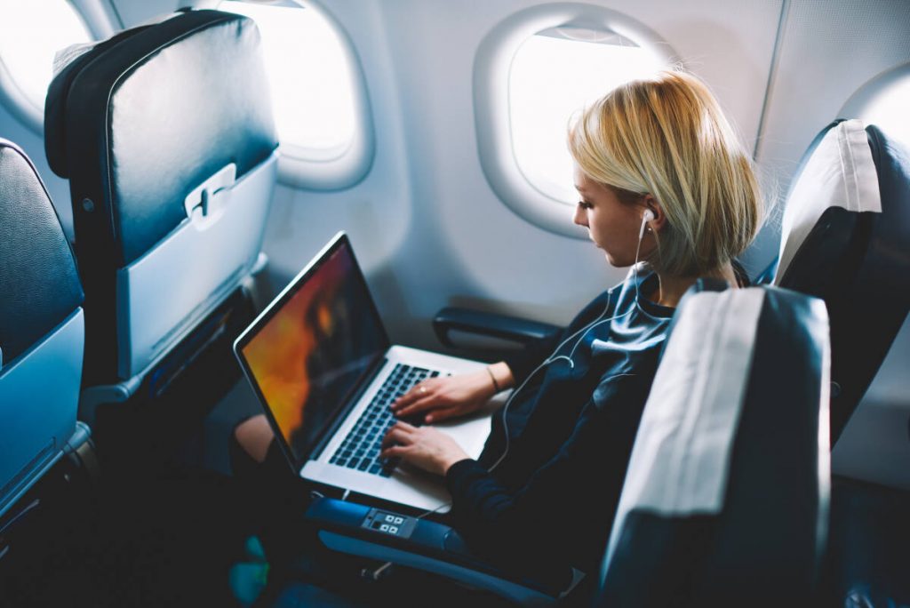 Business travel: A white female airplane passenger wearing a business suit uses a laptop computer while listening to headphones.