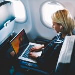 Business travel: A white female airplane passenger wearing a business suit uses a laptop computer while listening to headphones.