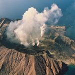 An aerial image shows a volcanic island emitting white smoke surrounded by clear blue sea on a clear day.