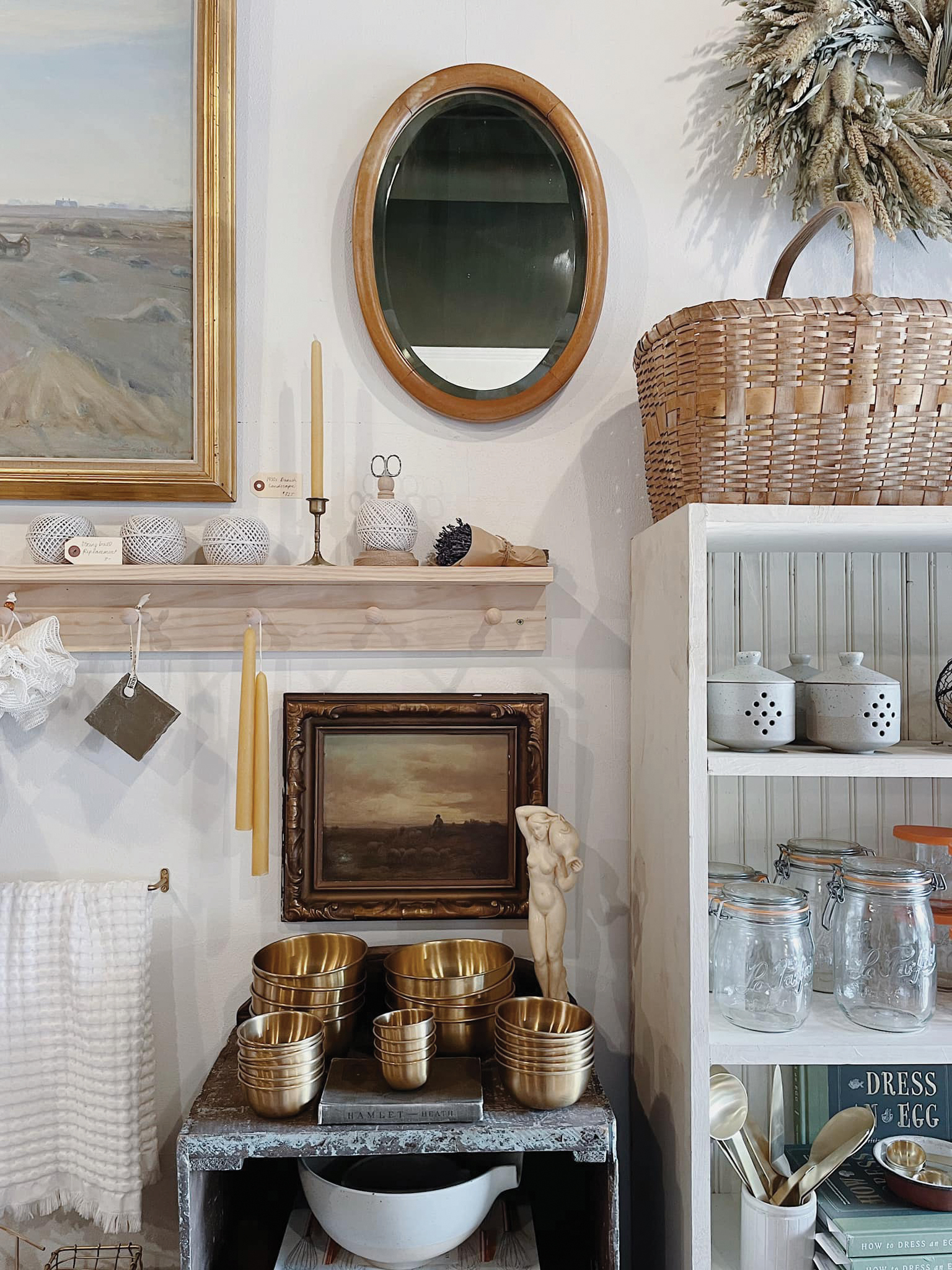Una toma de una tienda de artículos para el hogar con una mesa en el centro con ollas de cobre, una estantería blanca con cerámica y un estante encima con un candelabro, chucherías y un espejo.