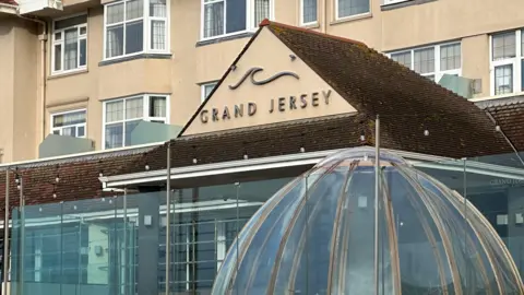 Frente al Grand Hotel en BBC Jersey. La fachada del edificio es gris con numerosas ventanas y el cartel está montado en la parte triangular del tejado. Debajo del cartel hay una estructura de vidrio con forma de cúpula.