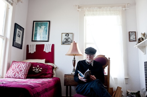 Un hombre lee un libro en una habitación de hotel con cuadros en la pared y una cama pequeña y luminosa.