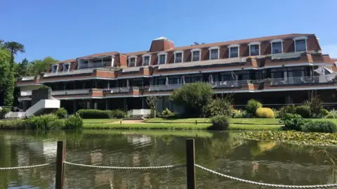 St. Pierre Park es un hotel de ladrillo rojo con balcones y frente al lago. Es un día de cielo azul y hay un poste y una valla metálica en primer plano.