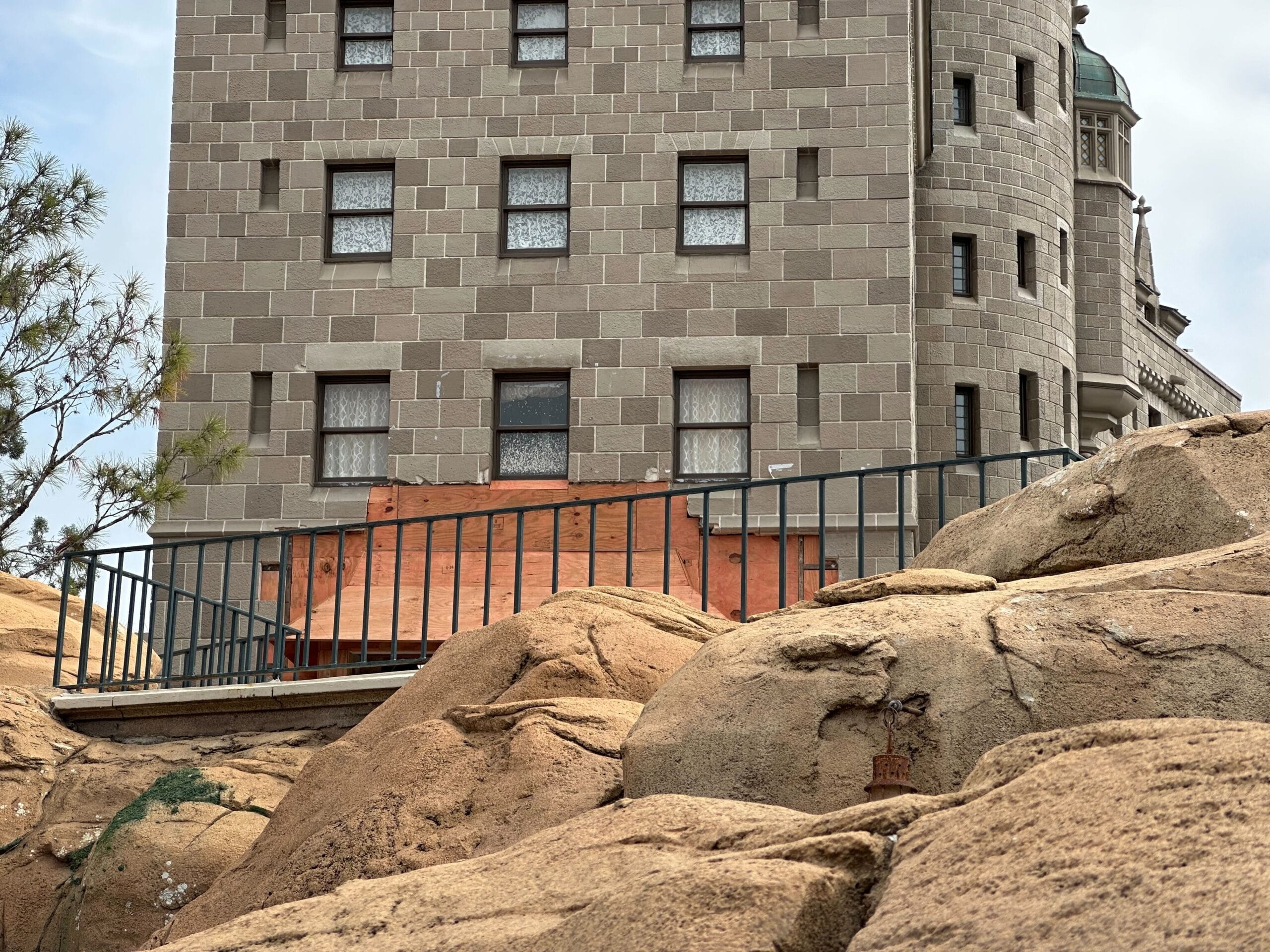 Edificio de piedra con muchas ventanas, bordeado por rejas metálicas y grandes rocas en primer plano.
