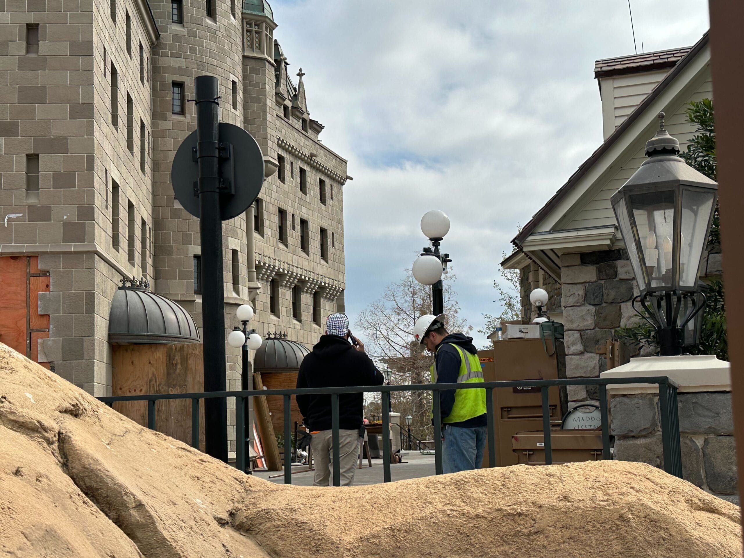 Dos trabajadores de la construcción con cascos discutiendo el trabajo parados junto a un edificio de piedra y una lámpara. La caja y el equipamiento son visibles al fondo.