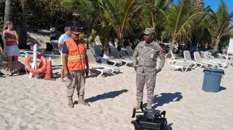 Los funcionarios de Defense Civil Dominica buscaron a Sudshaka Koni. Está rodeado de arena y sillas de playa vacías y un dron está en la arena cerca de sus pies. 