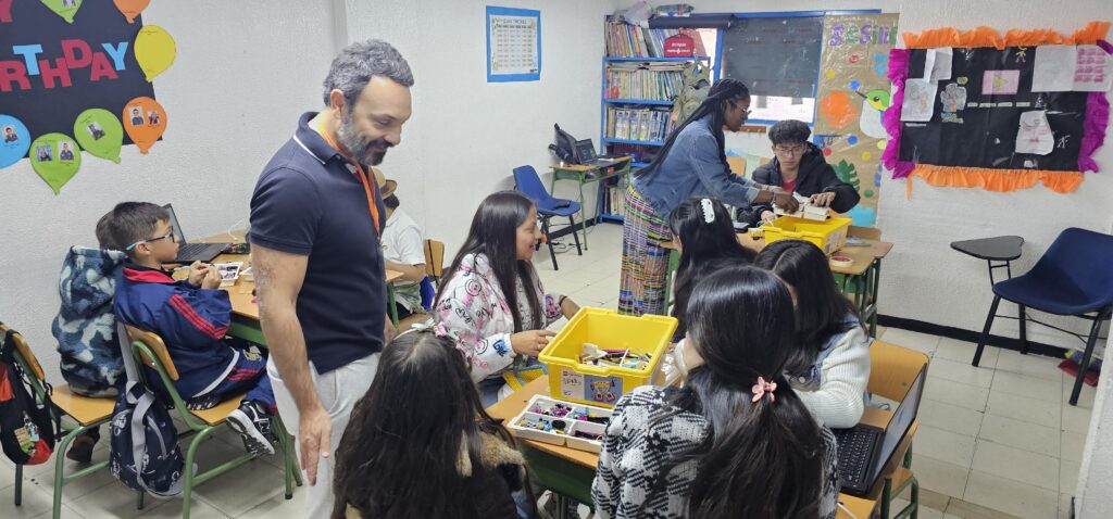 Los adultos y los niños interactúan alrededor de las mesas con bloques de construcción en el aula decoradas con carteles y estudiantes.