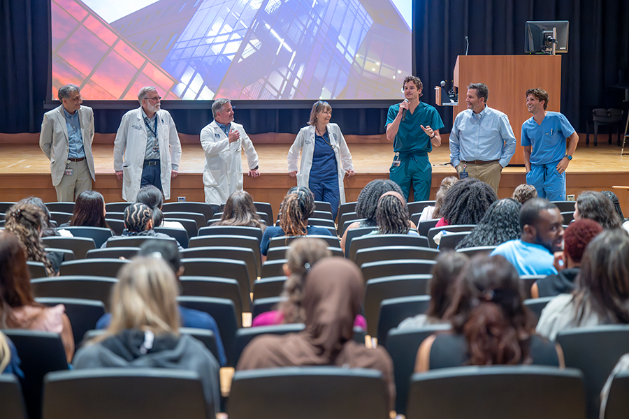 Siete profesores universitarios, incluidos seis hombres y una mujer, se paran frente al auditorio y hablan con un gran grupo de estudiantes universitarios.