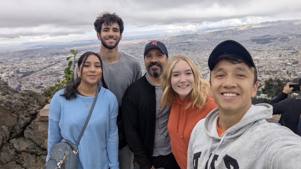 Cinco personas ofrecen un grupo de selfies en el fondo con miras de la ciudad y cielo nublado.