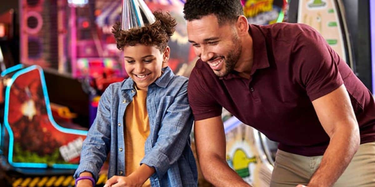 En el sombrero de fiesta, un bebé sonriente y una persona juegan un juego de arcade juntos, disfrutando de un arcade de entretenimiento de luz brillante.