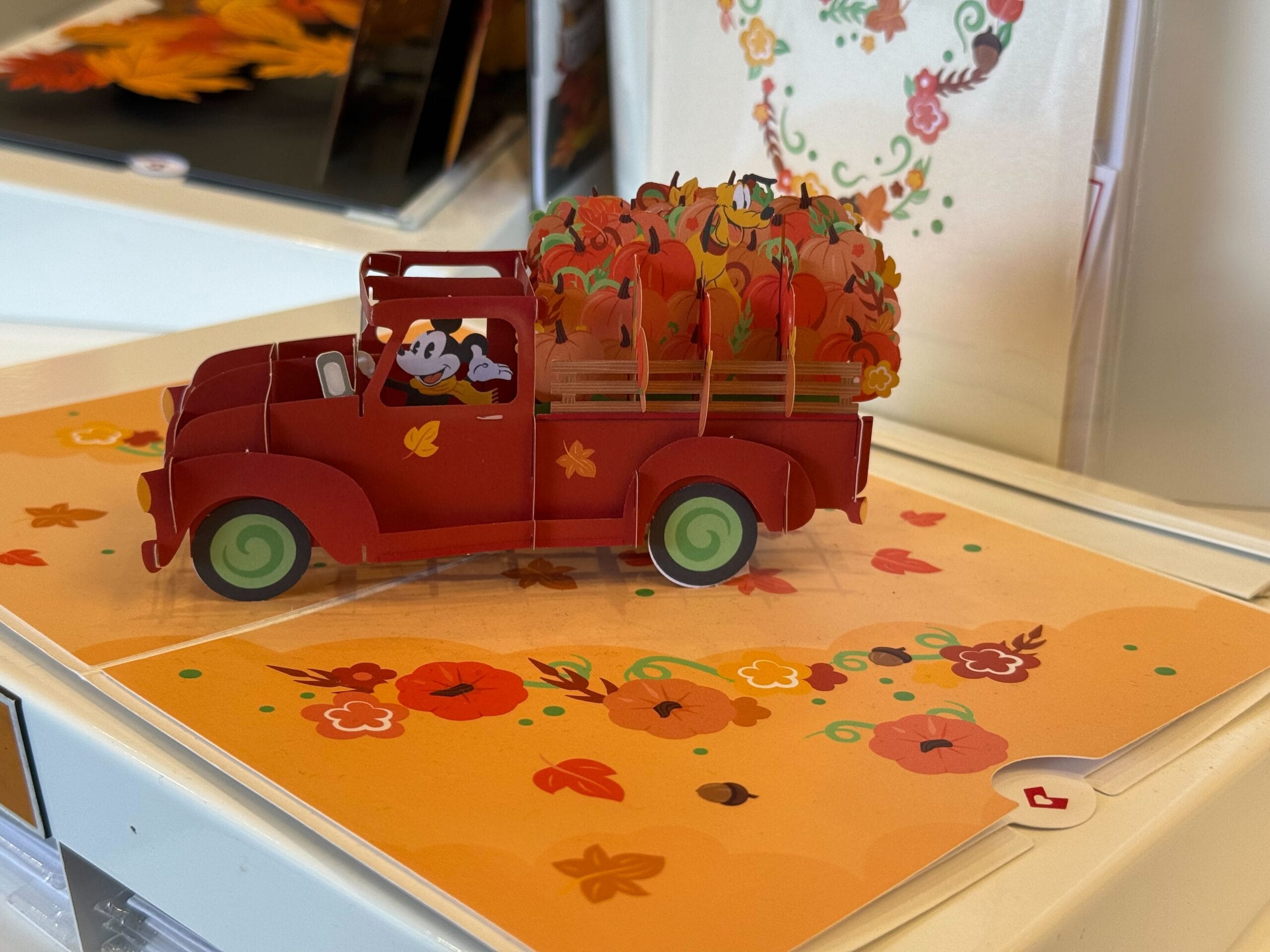 Una tarjeta de camión rojo -popa llena de calabaza se encuentra en la superficie naranja que está decorada con hojas y flores que caen.