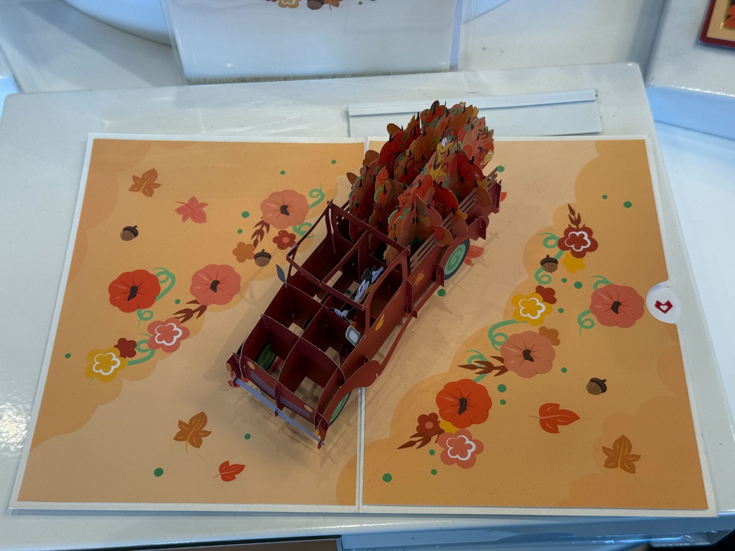 Una tarjeta emergente incluye un camión rojo 3D lleno de hojas de otoño y flores en el fondo de las flores de naranja.