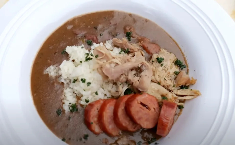 Un tazón de gumbo con pollo cortado, salchicha cortada y arroz, decorado con hierbas