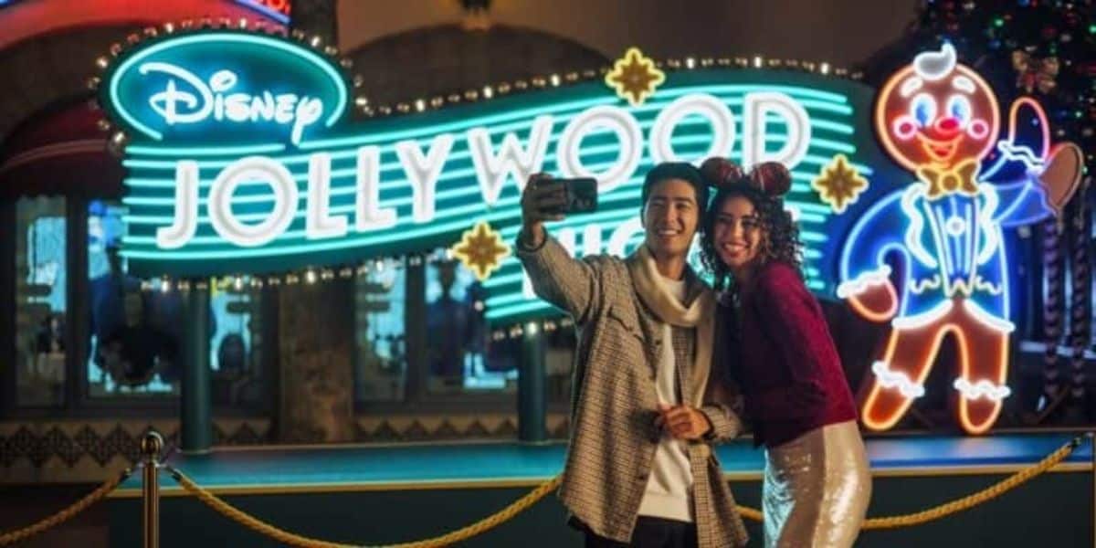 Por la noche, un hombre y una mujer se toman selfie frente a una brillante marca de madera de Disney Julie de Disney. Ambos son sonrientes y vistiendo ropa festiva, en la que la mujer toca la oreja mini ratón. Se puede ver un hombre de pan de jengibre de neón en el fondo.