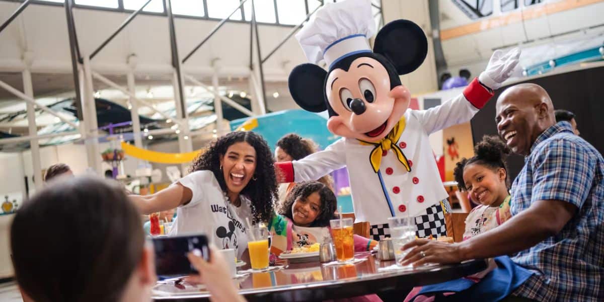 Una familia alegre con un hombre de cuatro veces vestido con el vestido de un chef en la mesa del comedor. Están disfrutando el servicio de mesa de comedor juntos, sobre la mesa con bebidas y platos de comida. Este diseño se ve en un lugar brillante de Disney cubierto.
