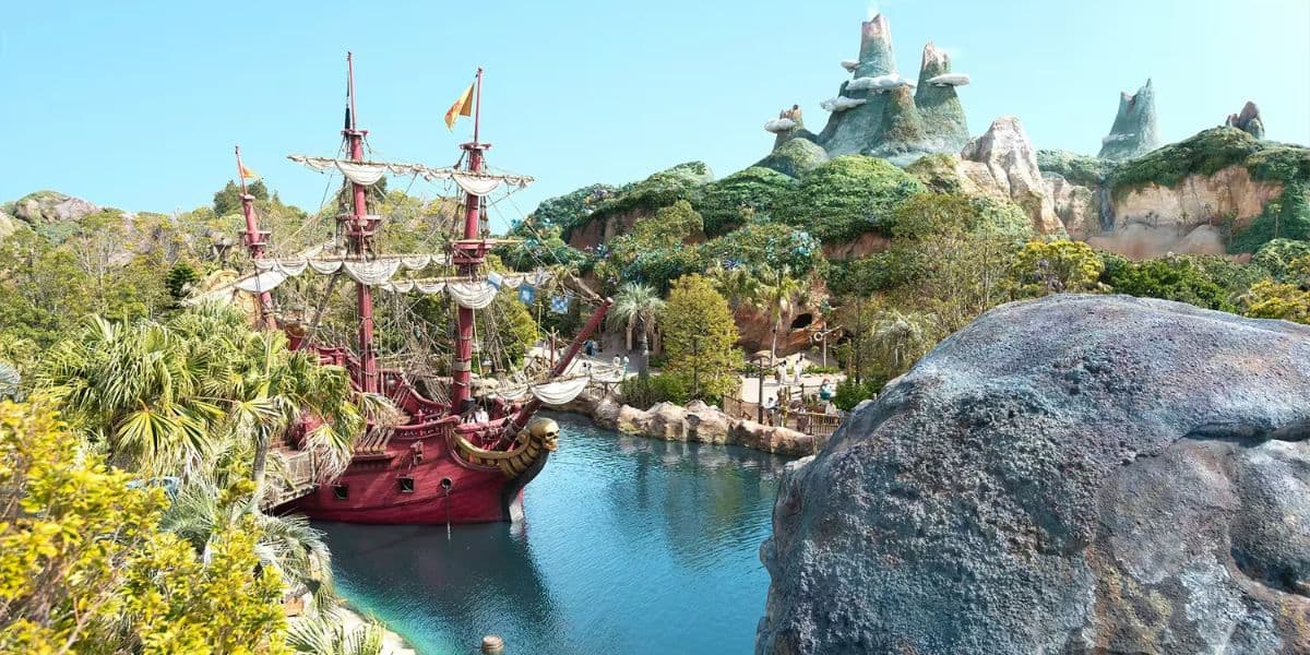 Una escena dinámica del parque temático con un barco pirata rojo en un canal de agua azul limpio rodeado de exuberantes plantas verdes. En el fondo, las emocionantes colinas rocosas se elevan bajo un cielo azul brillante.