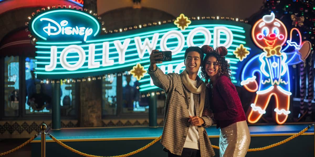 Una pareja feliz se toma una selfie frente al letrero brillantemente iluminado de Disney Jollywood. El hombre lleva una sudadera con capucha y la mujer lleva un vestido festivo con orejas de Minnie Mouse. Se destacan con decoraciones de muñecos de jengibre con luces de neón.