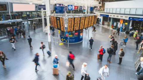 Getty Images Una vista frontal de la estación Manchester Piccadilly y el tablero central de salidas iluminado con destinos y horarios de trenes. Borrosas en la imagen debido al movimiento, una treintena de personas cruzan juntas.