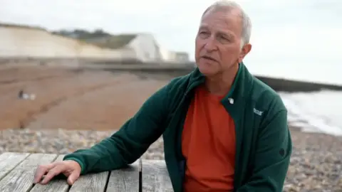 Foto de Giles Turner de Giles Turner con cabello blanco y vistiendo un top rojo y un vellón verde. Su mano descansa sobre una mesa. Al fondo se ven montañas, playa y mar. 