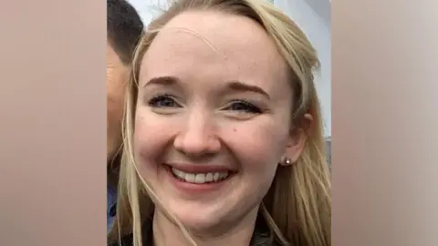 Andrea Brady Un primer plano de Jessica Brady sonriendo ampliamente y mirando directamente a la cámara, con cabello largo y rubio, ojos marrones y un arete en la oreja izquierda.
