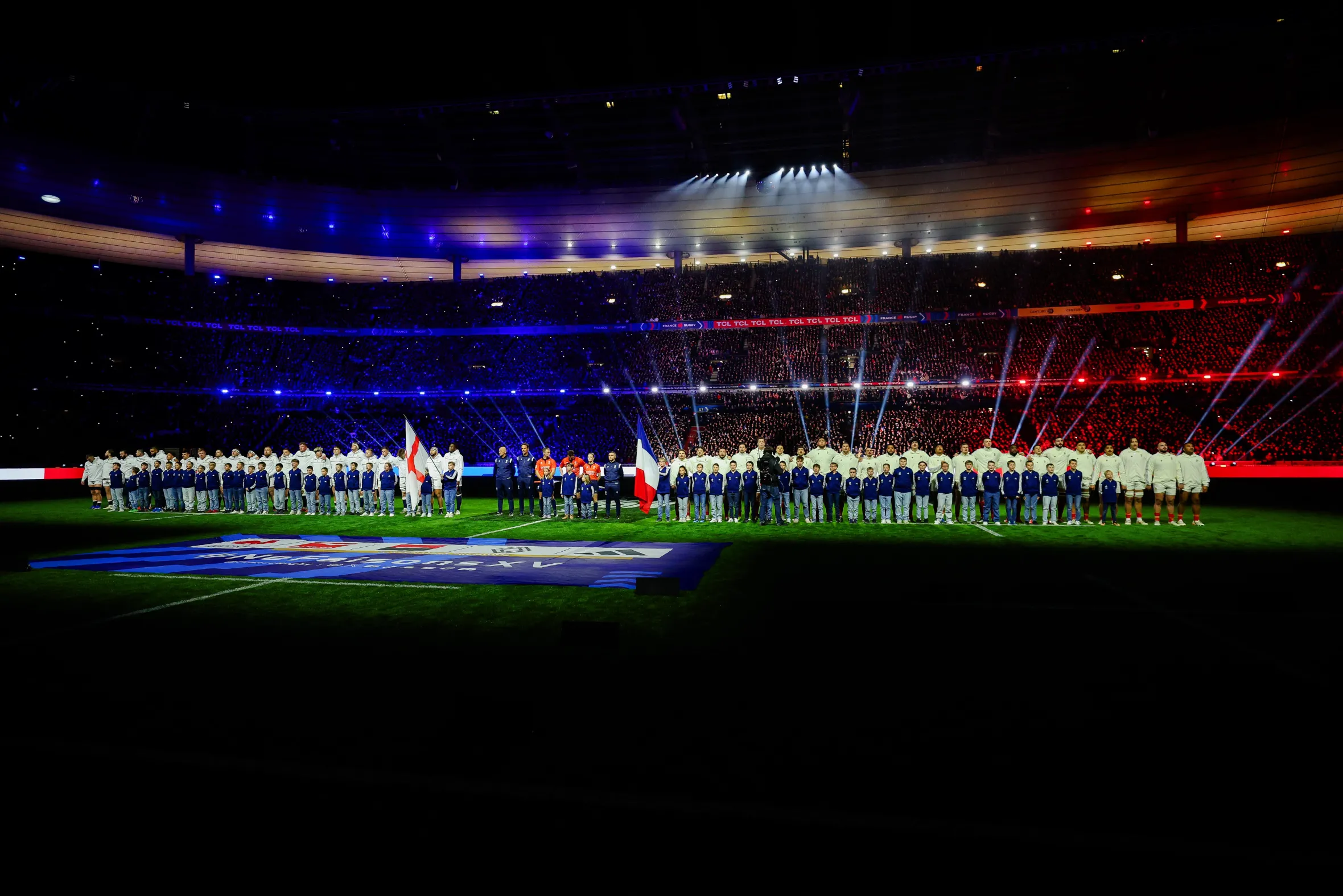 Los equipos de rugby y los niños se encuentran en el campo iluminado del estadio y los espectadores al fondo.