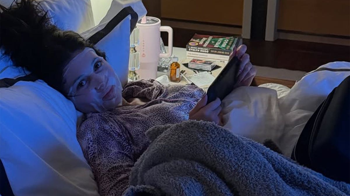 Kylie Jenner en una habitación de hotel oscura e iluminada de azul, vestida con un pijama con estampado animal y una mascarilla blanca. Ella sostiene un iPad y sonríe a la cámara. Al lado hay una mesa con bebidas y libros.
