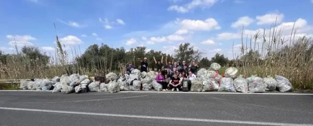 Un grupo de personas rodeadas de múltiples bolsas o basura al costado de la carretera, rodeadas de vegetación y cielos azules y despejados.