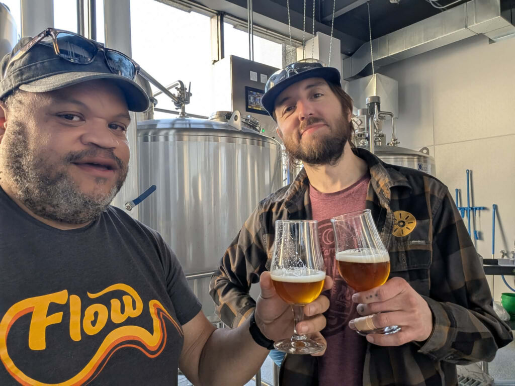 Dos hombres se encuentran dentro de una sala de cocción con vasos tipo tulipán de cerveza de color ámbar para brindar, con tanques de elaboración de cerveza de acero inoxidable y equipos al fondo.